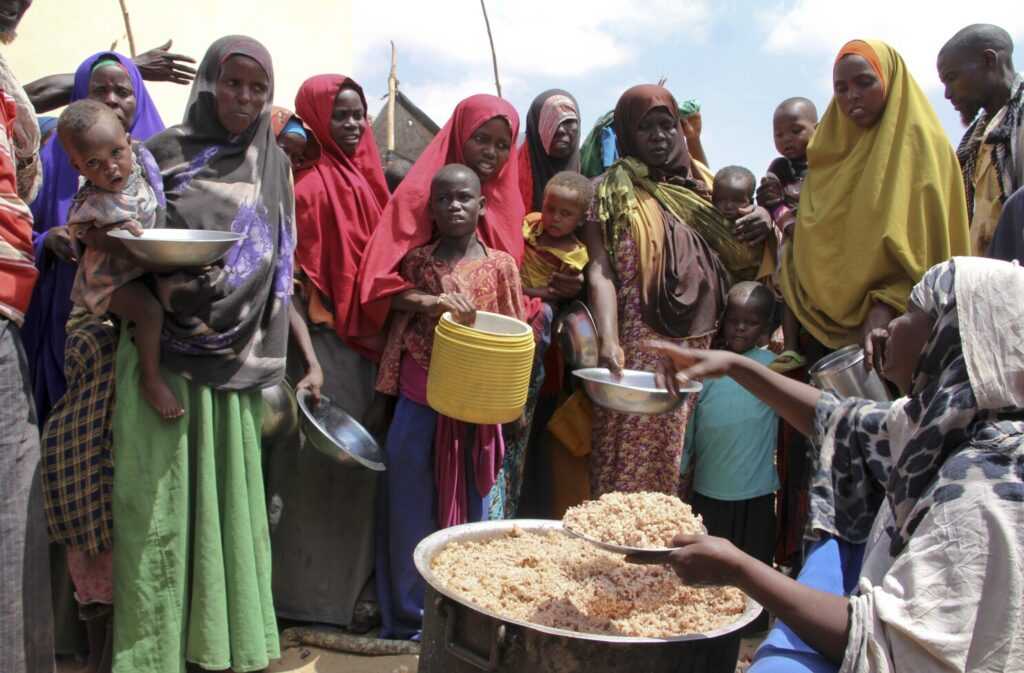 A group of African individuals carrying plates as they gather in line to receive food during a relief distribution, reflecting the severe impact of hunger and food insecurity.